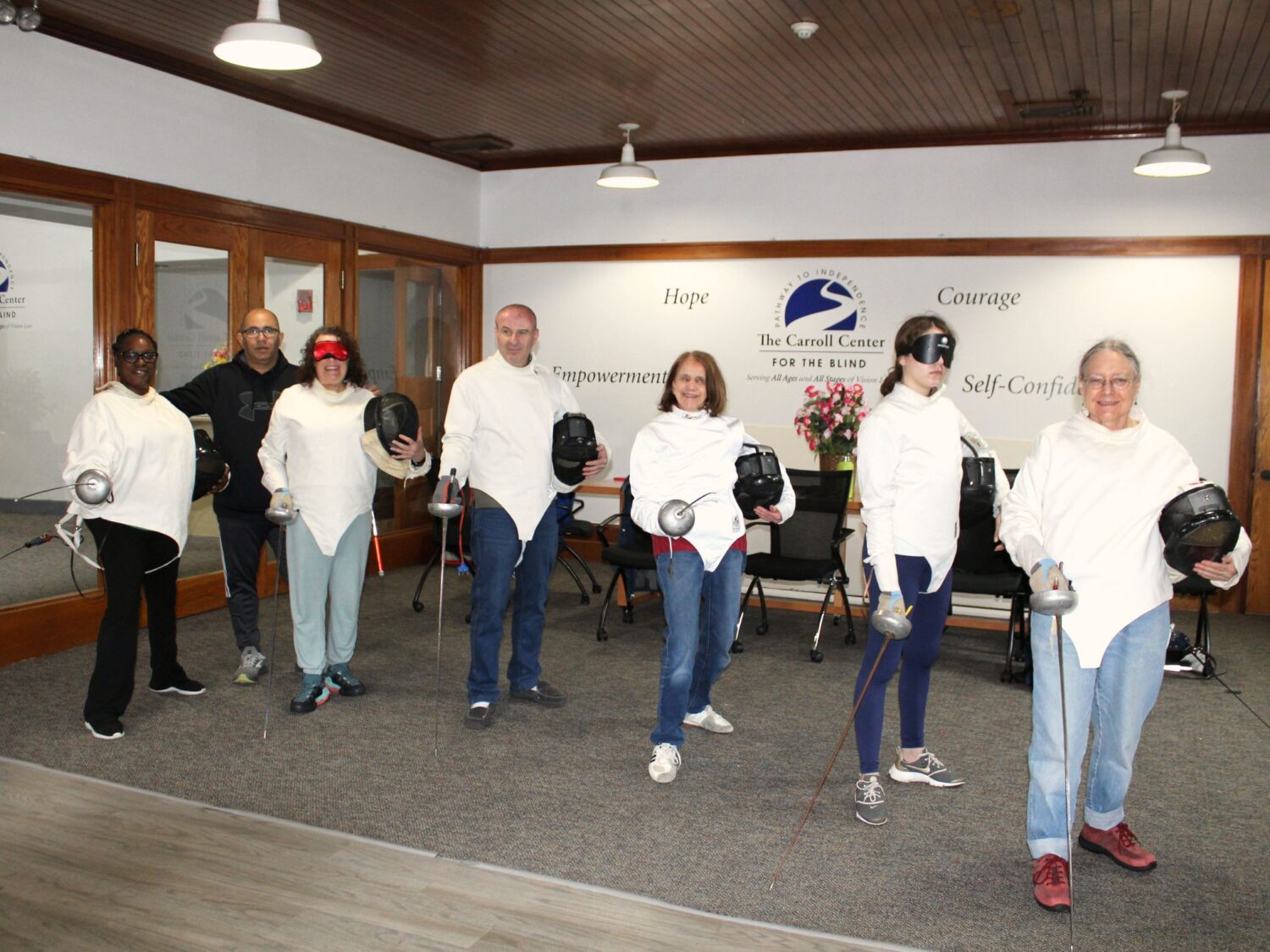 A group of 6 Carroll Center clients, in white fencing jackets holding masks and swords, pose together in a room. Their instructor is behind them. They are at The Carroll Center for the Blind, with words "Hope," "Courage," "Empowerment," and "Self-Confidence" visible on the wall behind them.