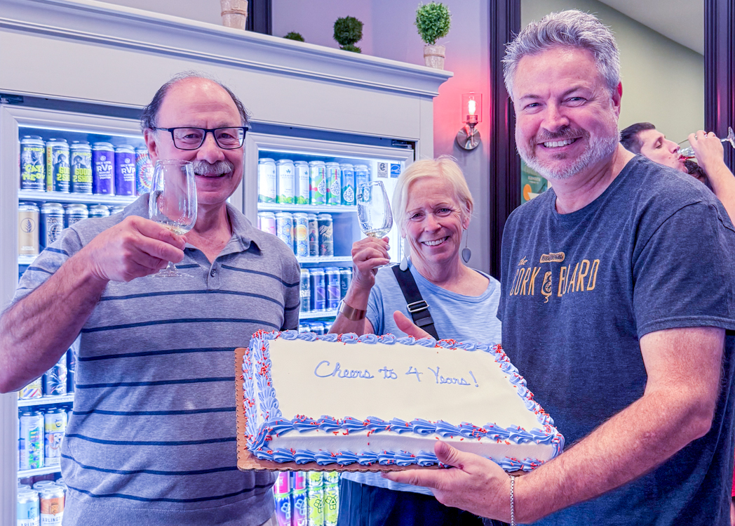 Ryan Gott holds a cake decorated "Cheers to 4 Years!" as two customers raise wine glasses in front of the store’s beer coolers